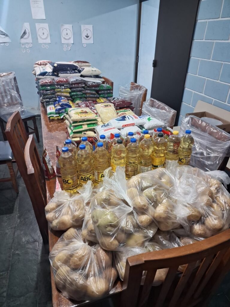 Salão do CEEB com os mantimentos separados sobre as mesas, arroz, feijão, óleo e sacos de batata, antes da montagem das cestas.